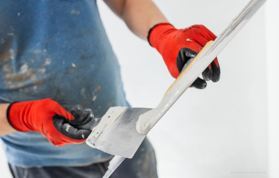Person mit roten Handschuhen, die mit einem Spachtel Trockenbaumasse auf eine weiße Oberfläche aufträgt.