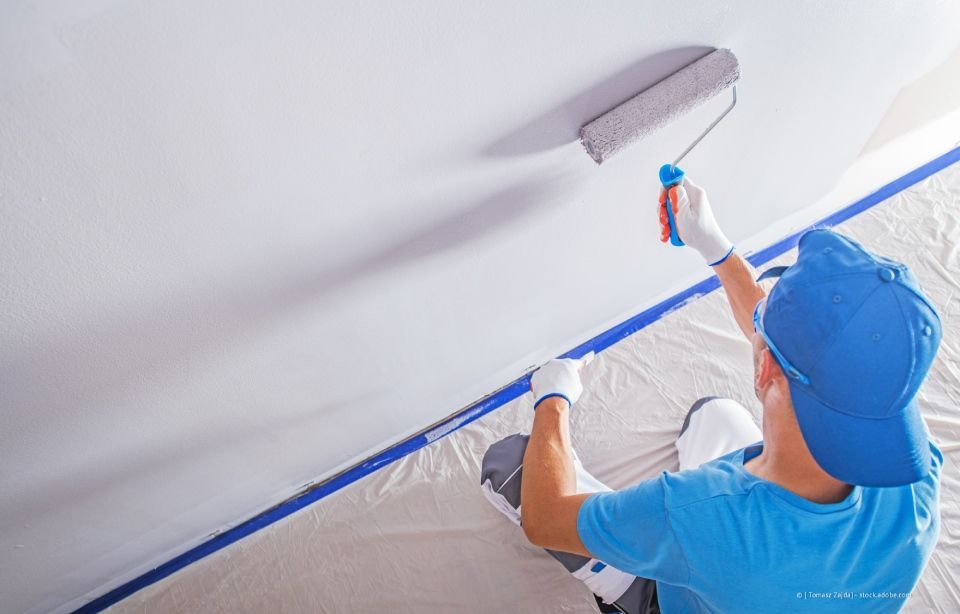 Person in Blau streicht eine weiße Decke mit einer Rolle und klebt die Ränder ab.