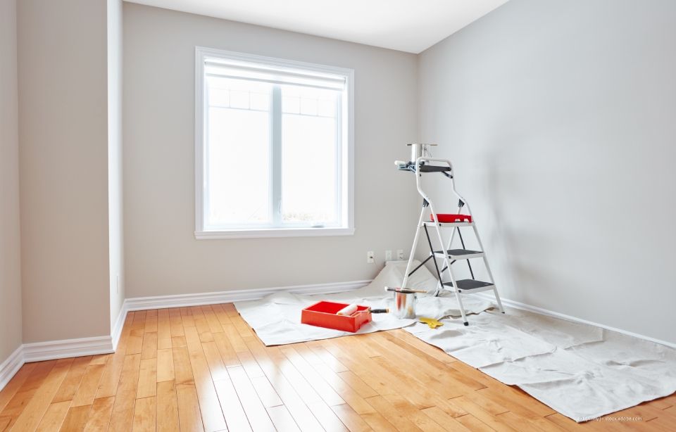 Der Raum wird gestrichen, mit hellgrauen Wänden, Hartholzböden und einem Fenster. Eine Leiter und Malerbedarf sind vorhanden.