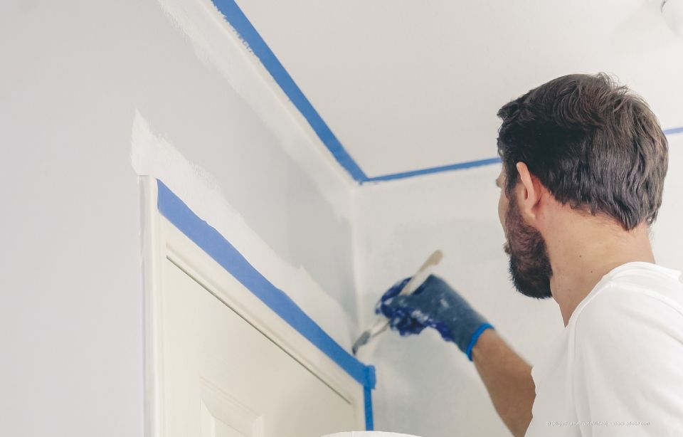 Mann, der ein weißes Hemd und blaue Handschuhe trägt und mit blauem Klebeband eine weiße Wand bemalt.