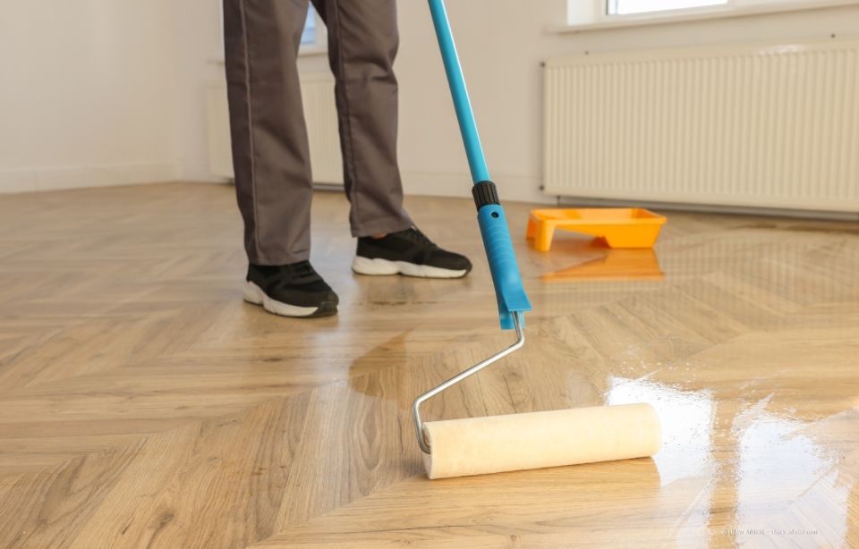 Person, die im Innenbereich mit einer Rolle Versiegelung auf einen Holzparkettboden aufträgt.