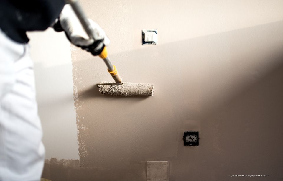 Person streicht eine Wand mit einer Farbrolle; die Wand ist beige und die Person trägt einen weißen Overall.