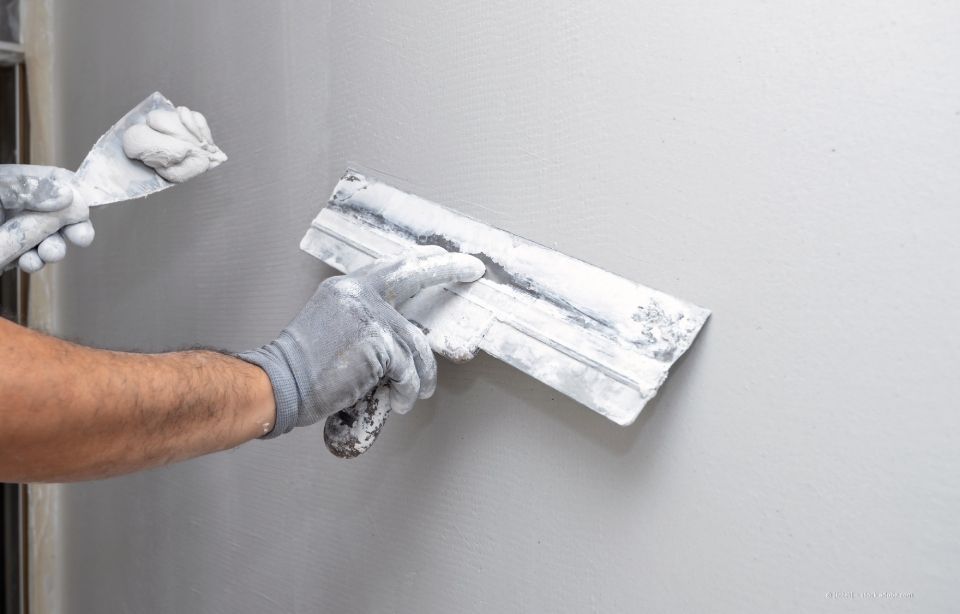 Person, die mit einem Spachtel weißen Spachtel auf einer grauen Wand glättet.