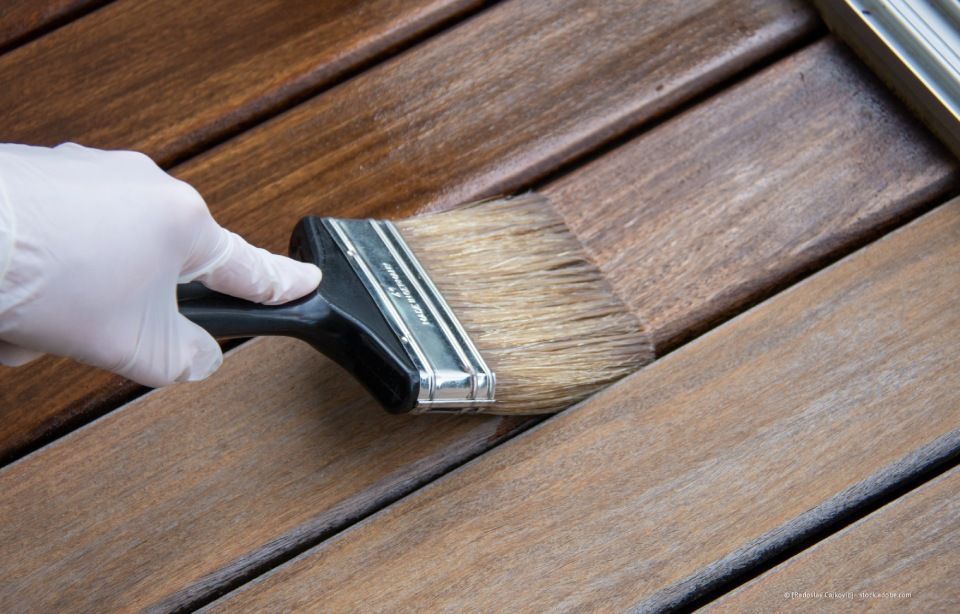 Behandschuhte Hand trägt mit einem Pinsel braune Holzbeize auf Holzbretter auf.