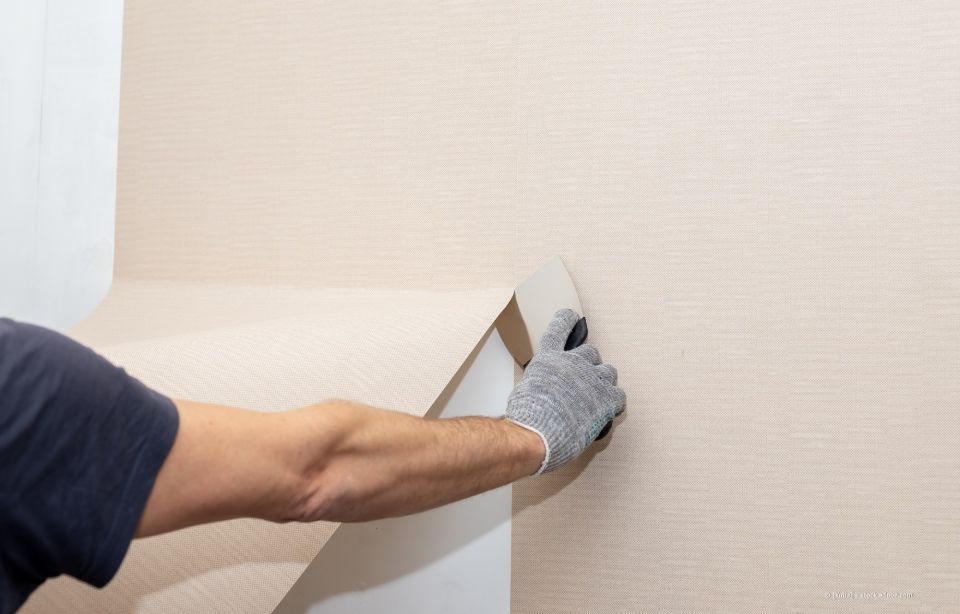 Person, die hellbeige Tapeten von einer Wand abzieht und einen grauen Handschuh trägt.