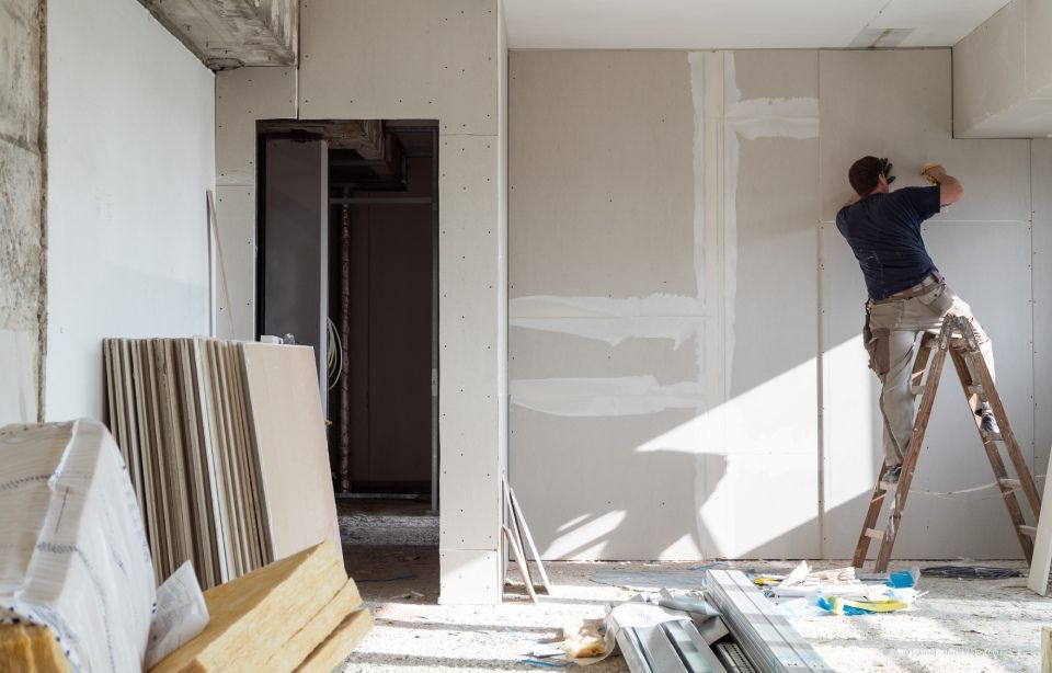 Mann auf einer Leiter installiert Trockenbauwände in einem im Bau befindlichen Raum; Sonnenlicht strömt herein.