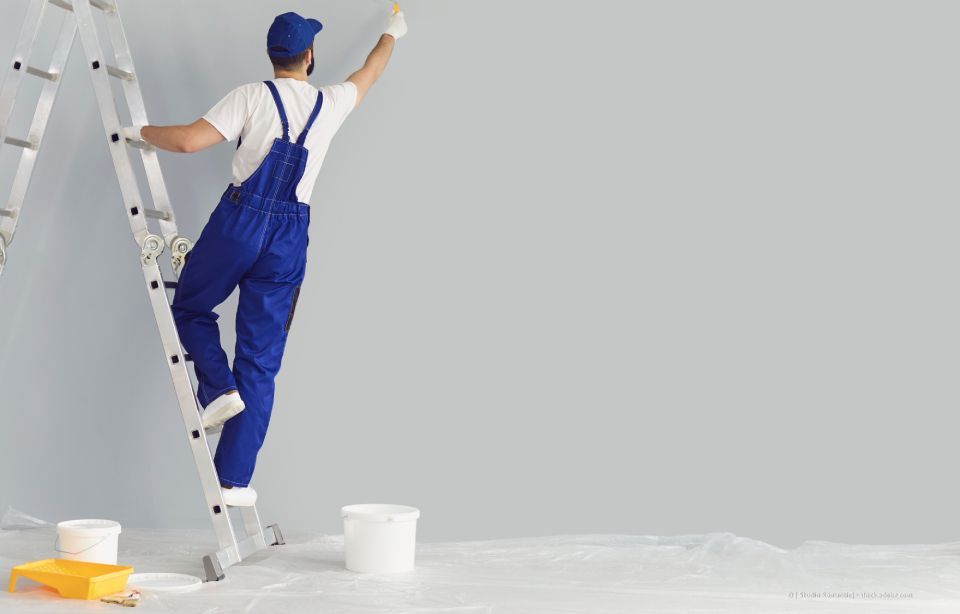 Maler im blauen Overall auf einer Leiter, der nach oben greift, um eine graue Wand zu streichen.