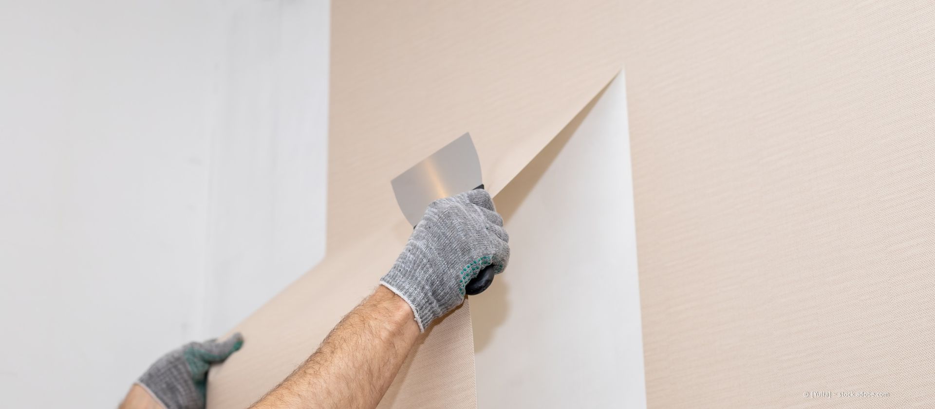 Eine Person entfernt mit einem Schaber Tapeten von einer Wand. Sie trägt Handschuhe.