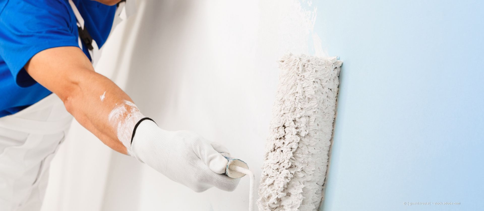 Eine Person streicht mit einer Farbrolle eine Wand blau. Sie trägt Handschuhe und ein blaues Hemd.