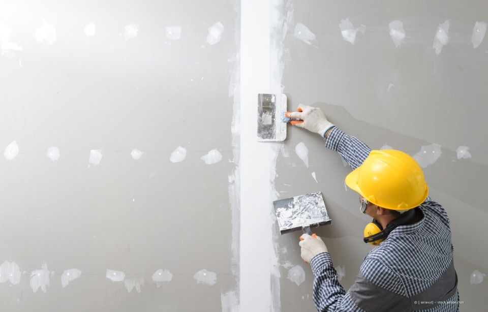Person mit gelbem Schutzhelm, Atemschutzmaske und Handschuhen glättet mit einer Kelle Trockenbaumasse an einer Wand.