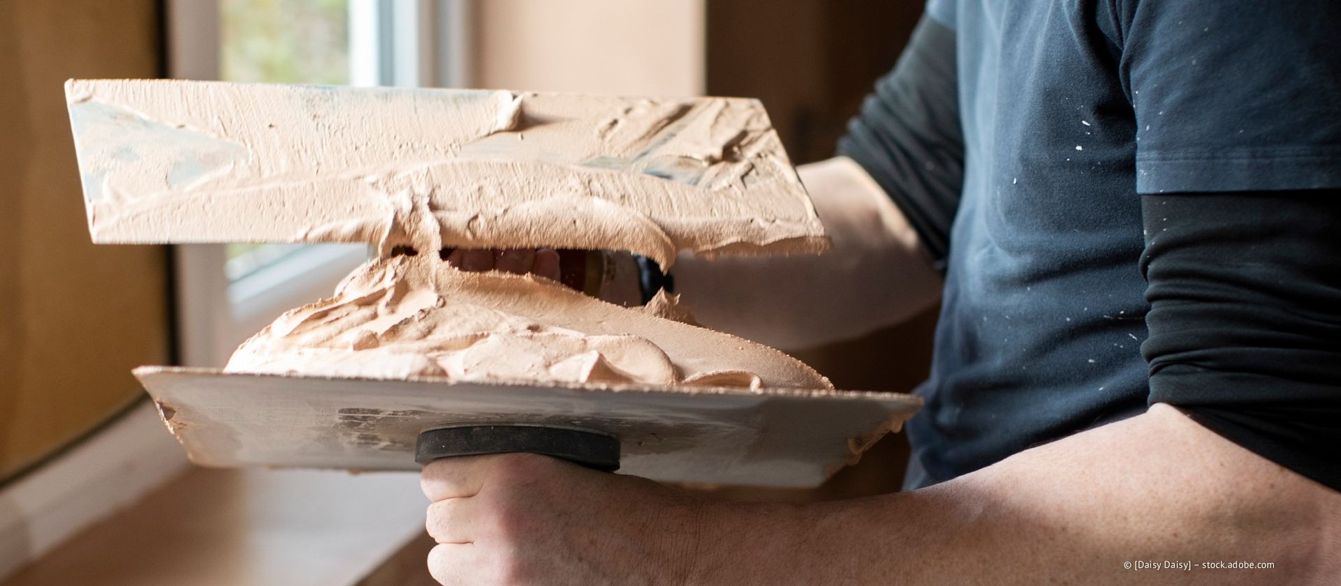 Person hält eine Kelle mit dickem, hellbraunem Gips, wahrscheinlich für Wandarbeiten.