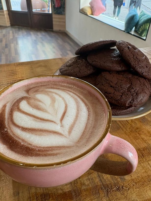 Una taza de café con leche rosa con un dibujo de corazón junto a una galleta de chocolate sobre una mesa de madera.