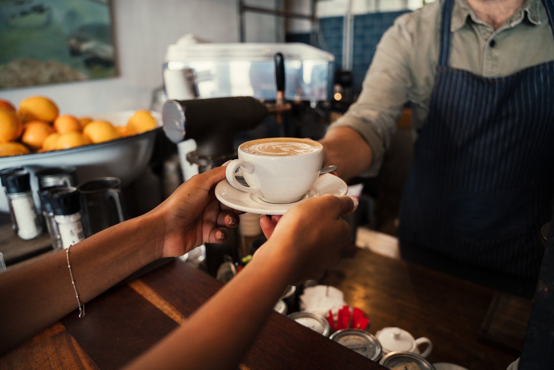 Un barista entrega una taza de capuchino a un cliente sobre un mostrador de madera en una cafetería.