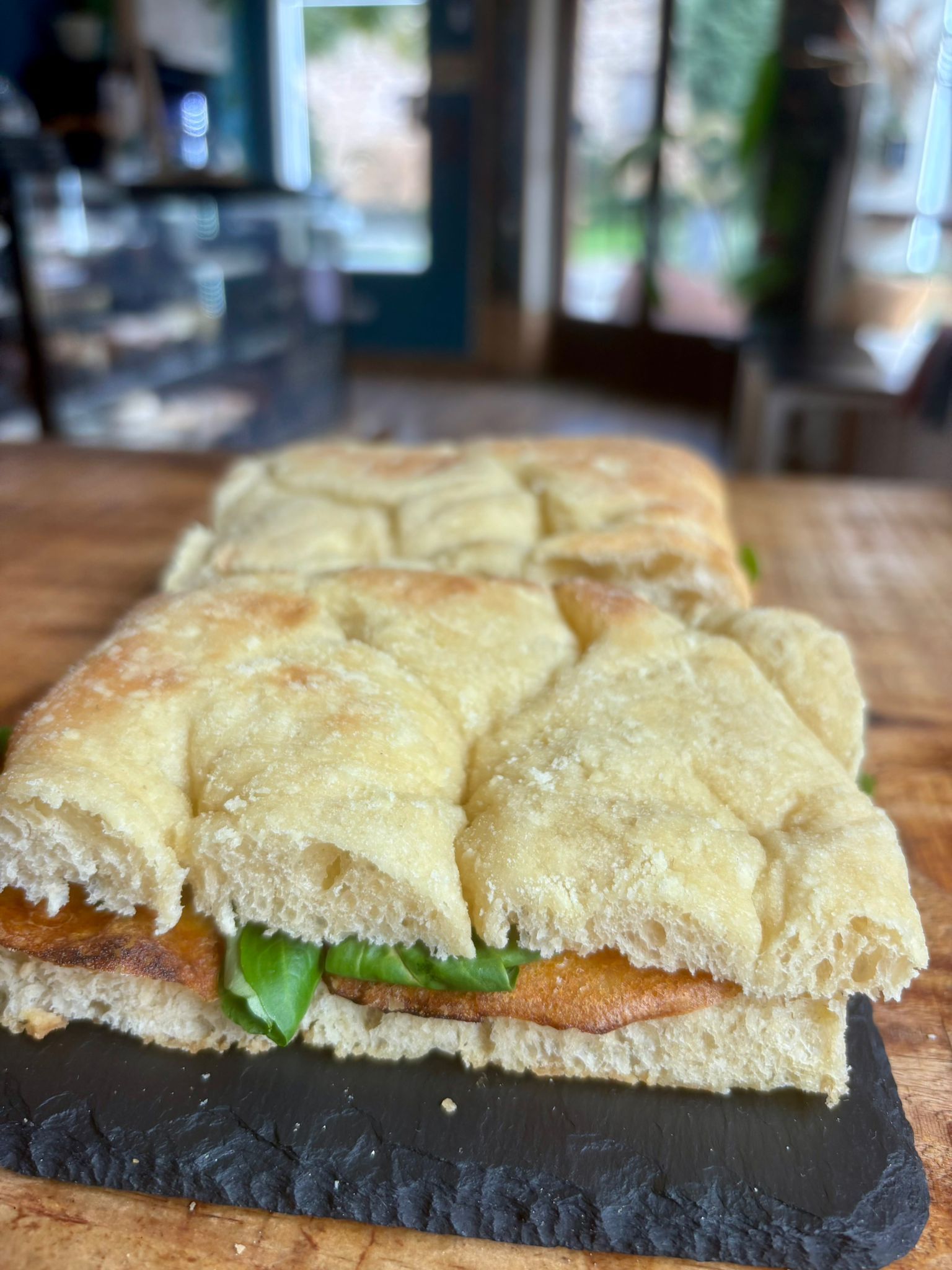 Sándwich de focaccia con tomate y verduras sobre una pizarra oscura en un entorno de cafetería.