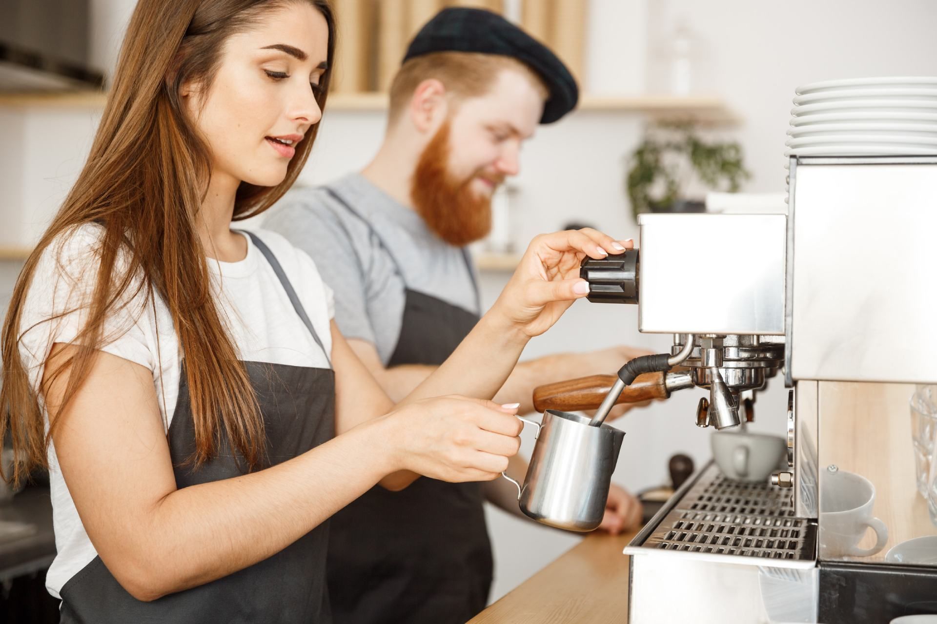 Una mujer calienta leche usando una máquina de espresso. Un hombre al fondo. Ambos llevan delantales en una cafetería.