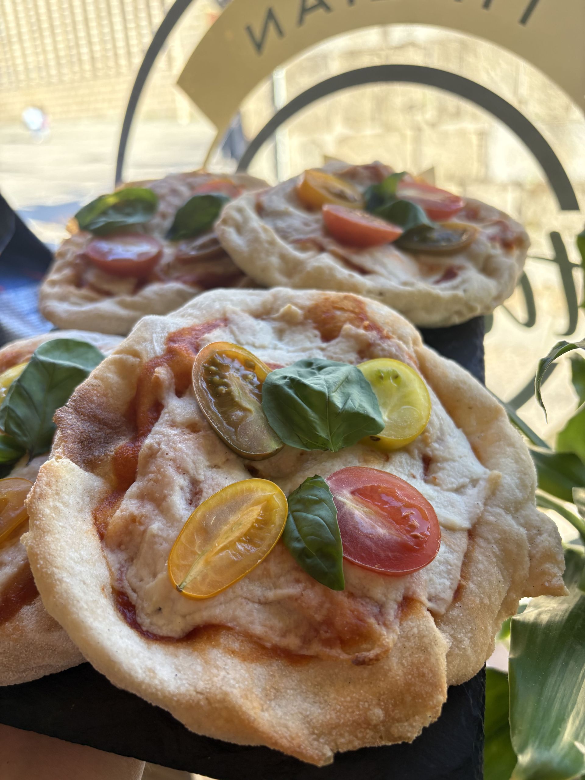 Mini pizzas cubiertas con tomates y albahaca en un plato negro, cerca de una ventana.