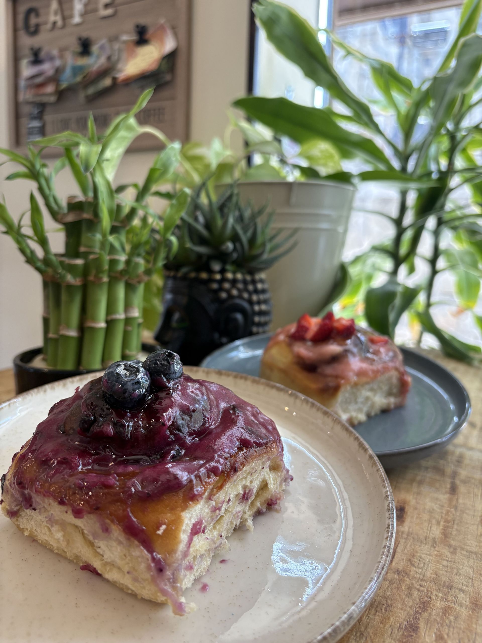 Dos bollos glaseados con arándanos y fresas, en platos, en un ambiente de cafetería con plantas.