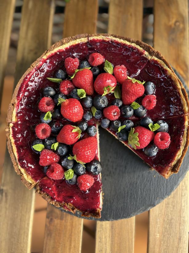 Tarta cubierta de frutos rojos, con una porción retirada, sobre un soporte redondo negro encima de una mesa de madera.
