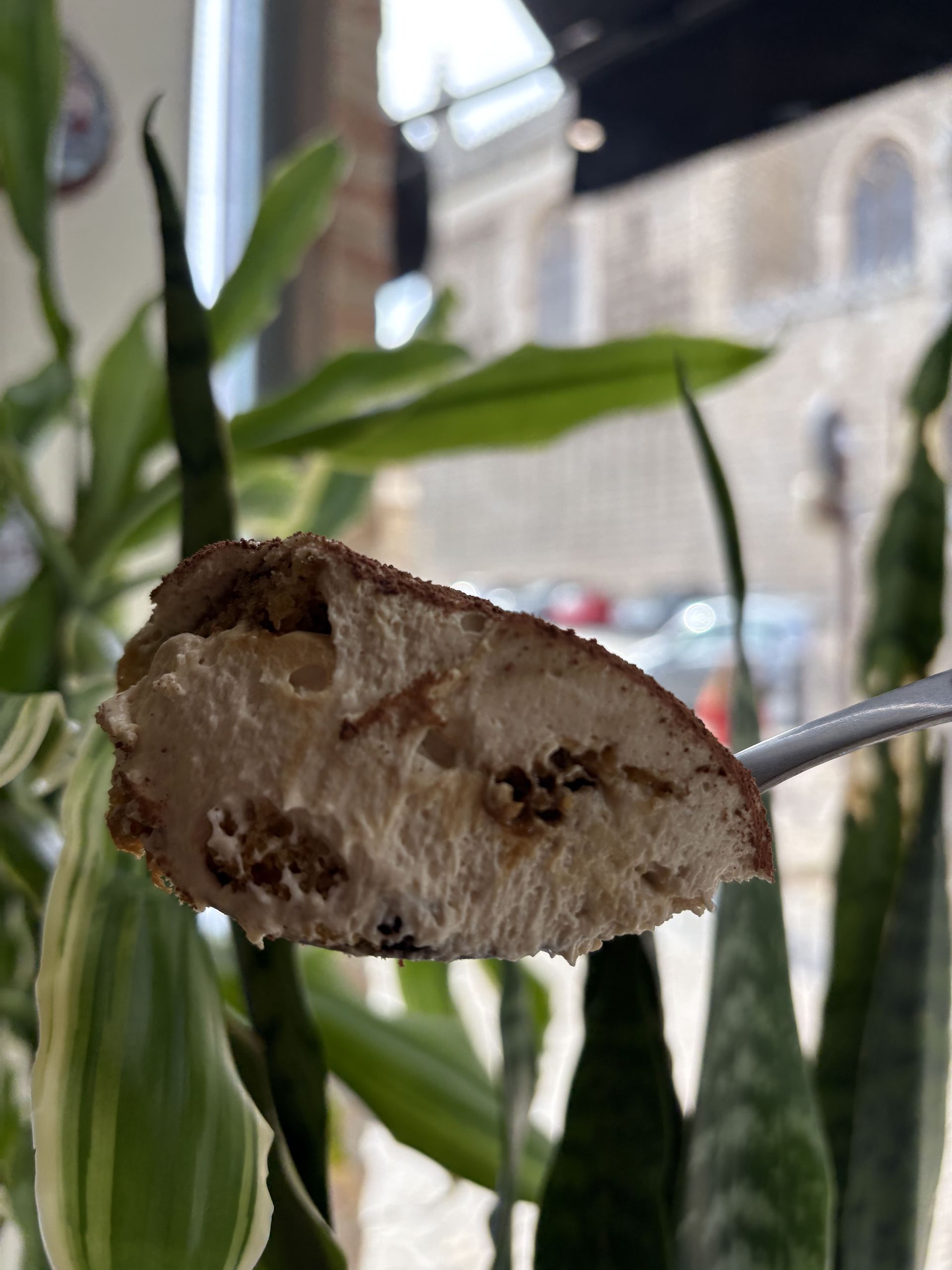 Una cucharada de tiramisú con capas visibles, sostenida contra un fondo borroso de un edificio y vegetación.