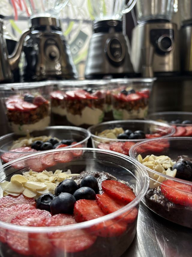 Vasitos de postre decorados con fresas, arándanos y crema sobre una mesa de exposición con máquinas de café detrás.