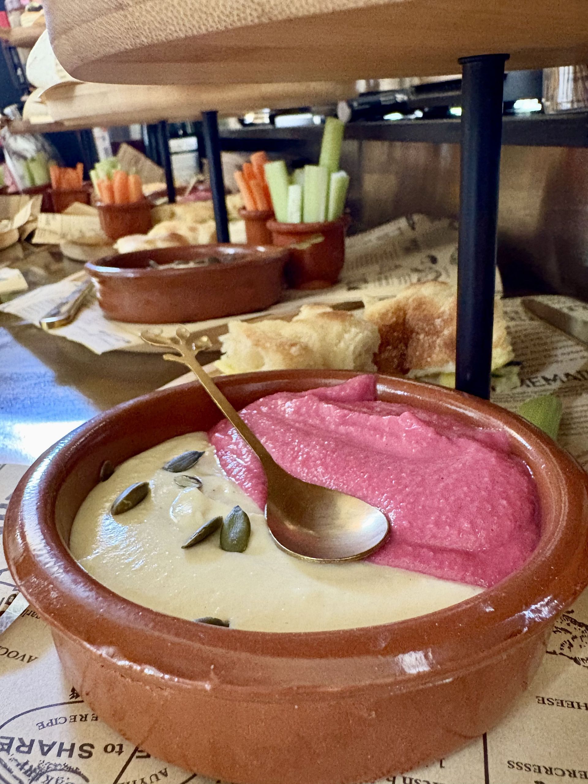 Una pequeña fuente de barro con hummus bicolor, pipas de calabaza y una cuchara dorada. Verduras al fondo.