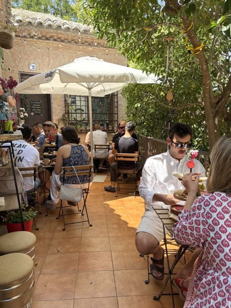 Patio de restaurante al aire libre con clientes en mesas bajo una sombrilla, cerca de árboles y un edificio.