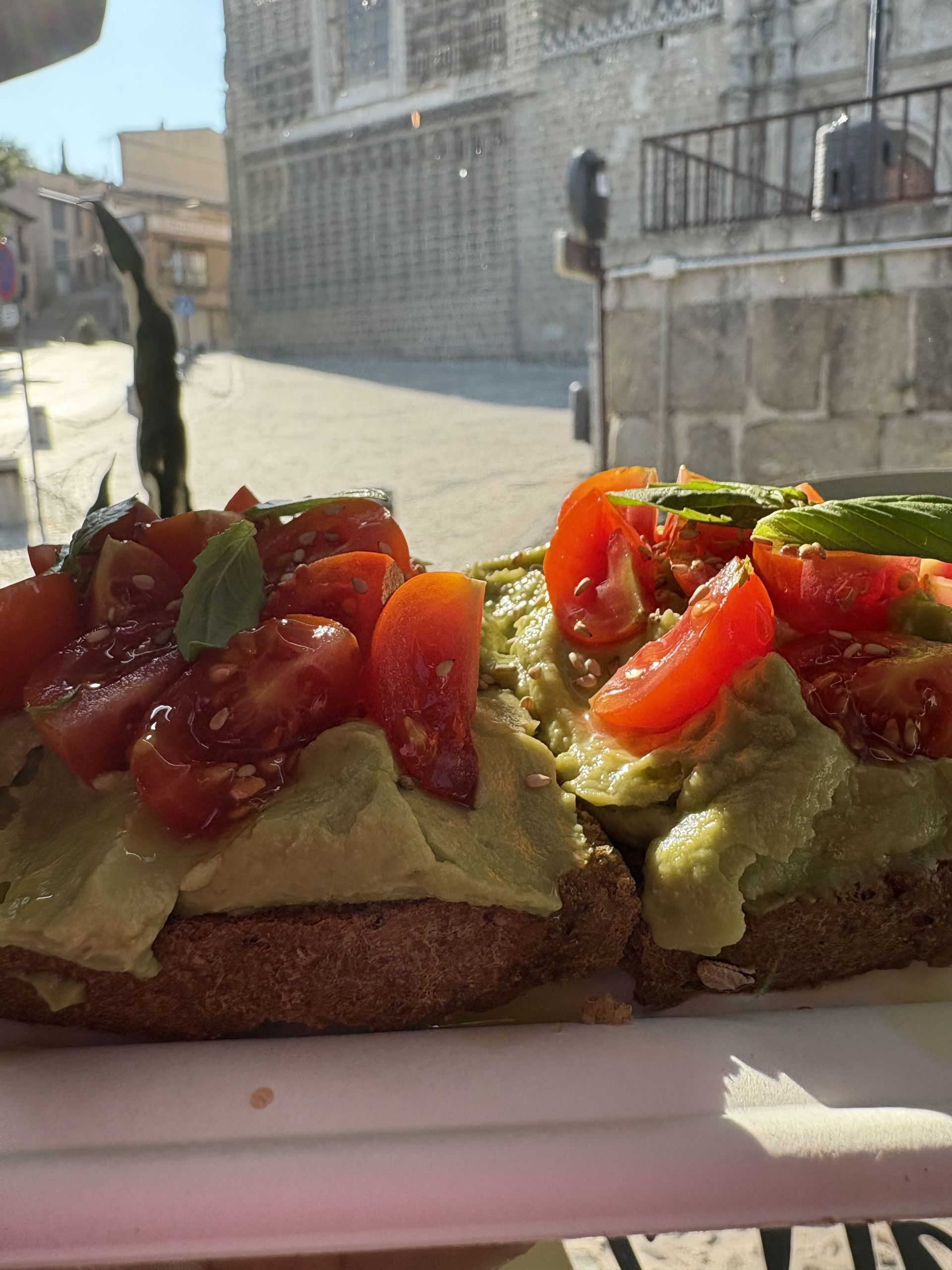 Tostada de aguacate cubierta con tomates en rodajas, al aire libre y con un edificio de piedra como fondo.