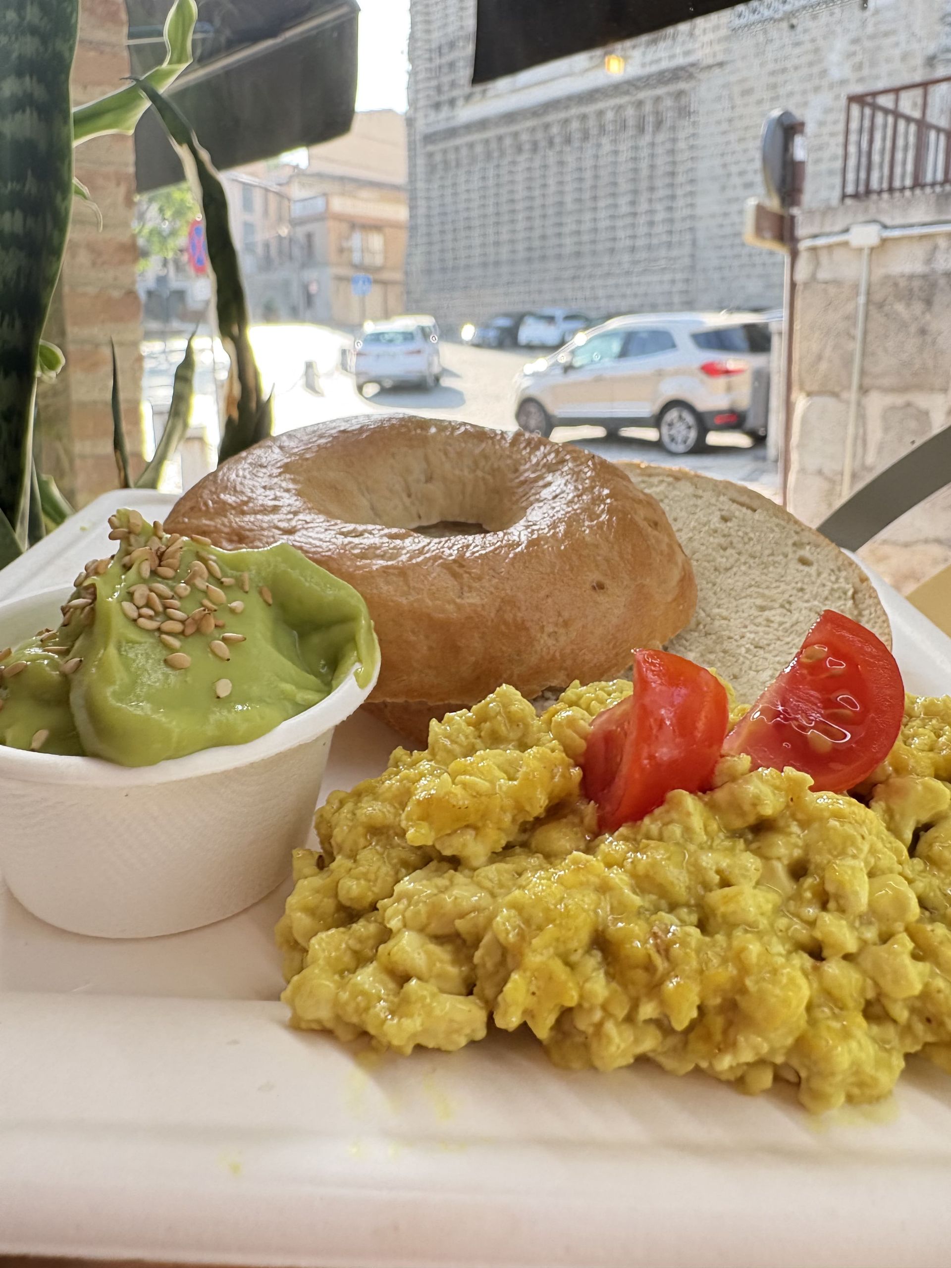 Plato de desayuno con huevos revueltos, bagel, aguacate y tomates; fondo de calle.