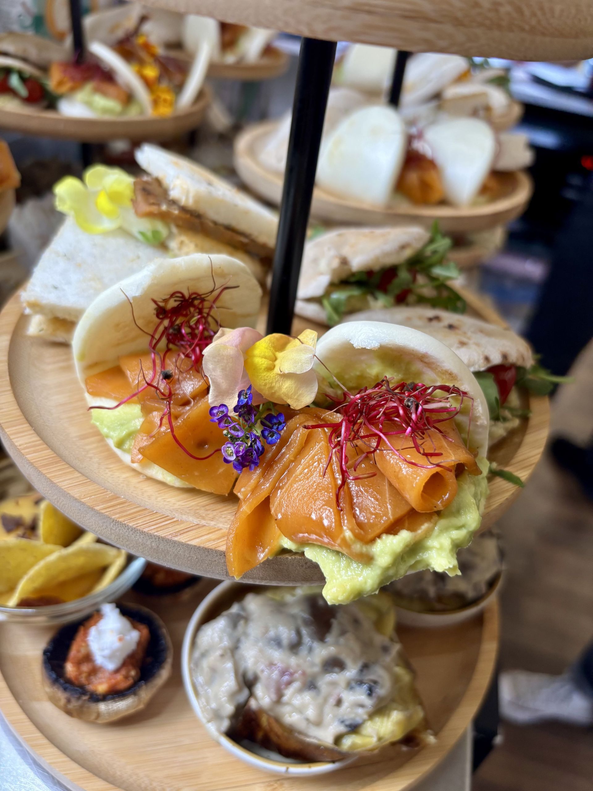 Bandeja de madera escalonada para servir con aperitivos variados: bollos de salmón, sándwiches y tartas pequeñas.