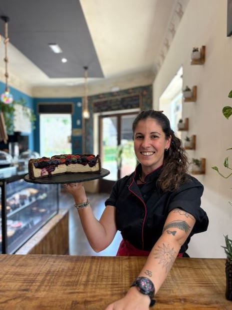 Mujer con chaqueta de chef negra sosteniendo una tarta de queso cubierta de bayas en una cafetería.