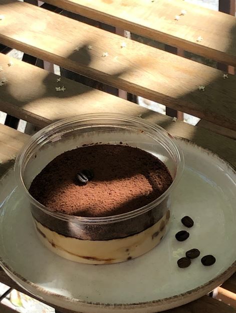 Postre de tiramisú en un recipiente de plástico transparente sobre un plato blanco con granos de café.