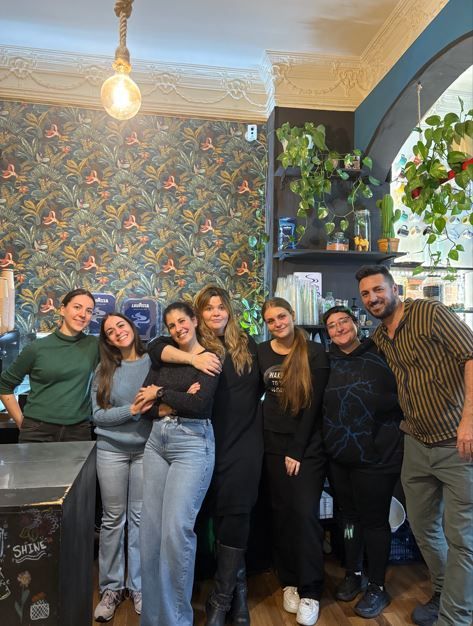 Grupo de personas sonriendo juntas dentro de un café, con papel tapiz floral y plantas.