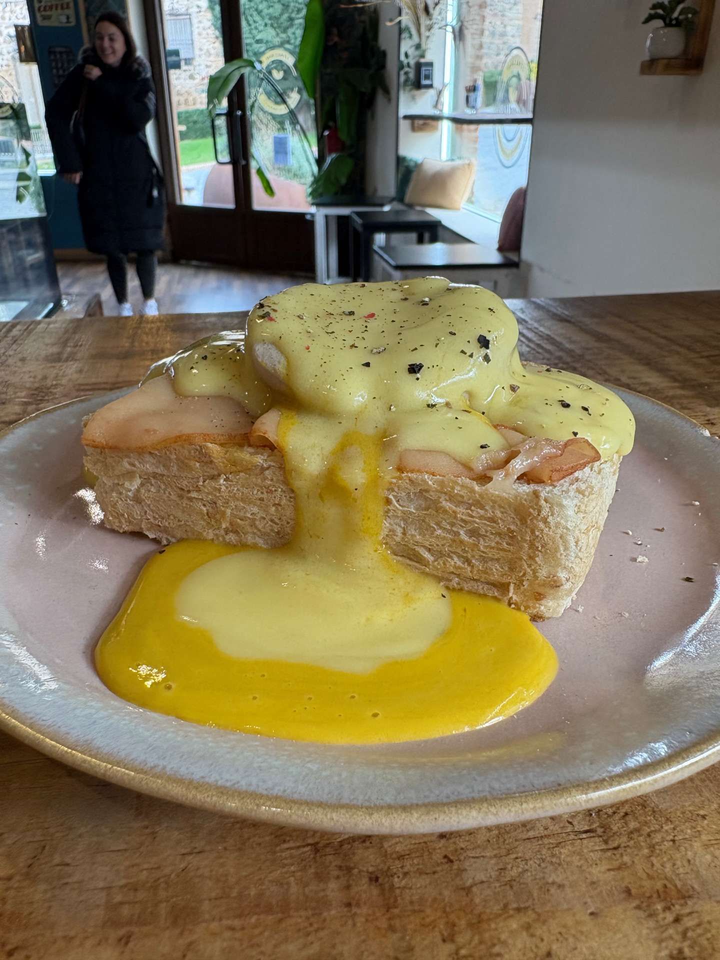 Huevos Benedict sobre tostada, con salsa holandesa amarilla derramándose sobre un plato rosa. Una persona de pie al fondo.