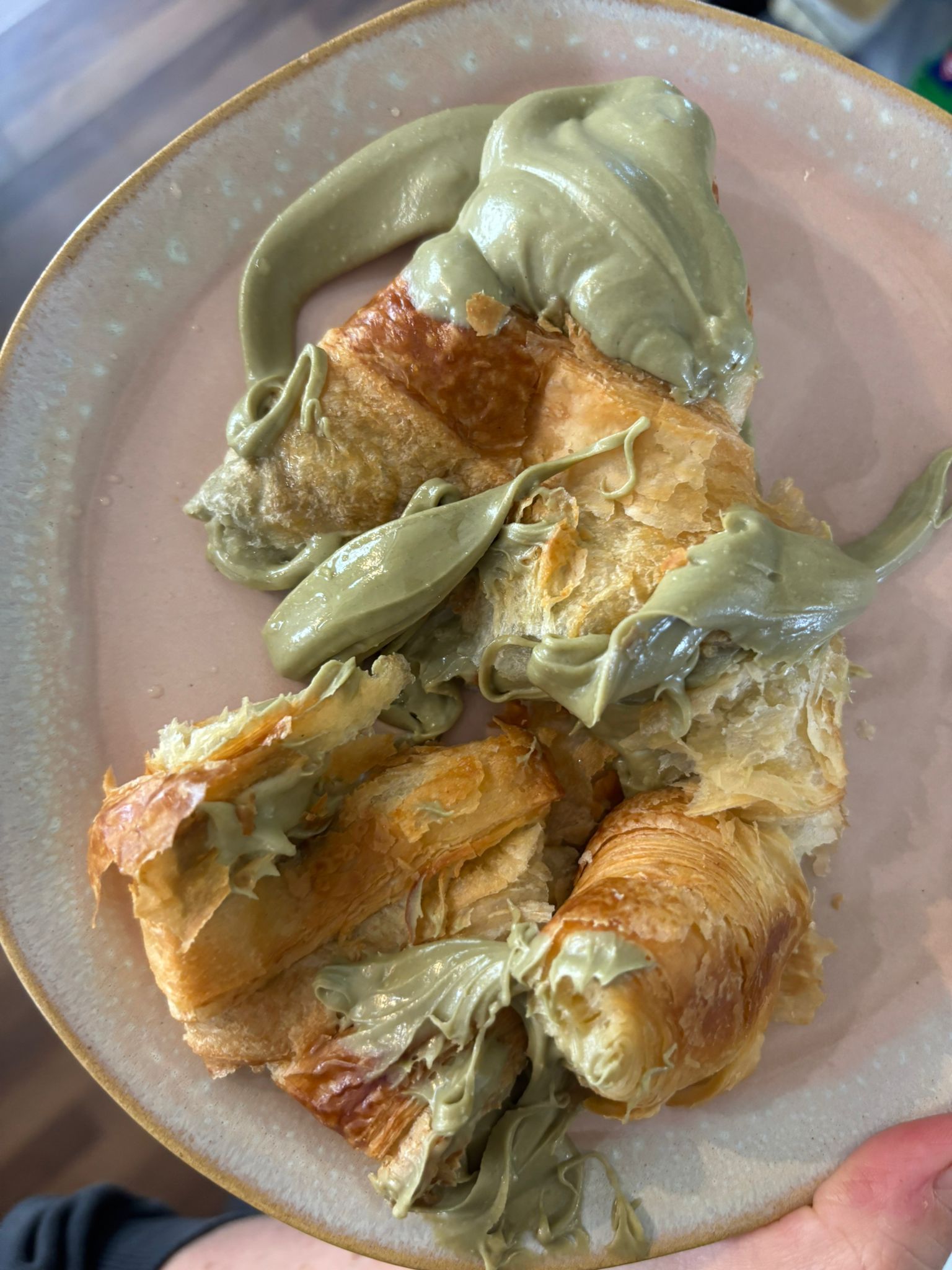 Croissant rociado con crema de pistacho verde sobre un plato rosa.