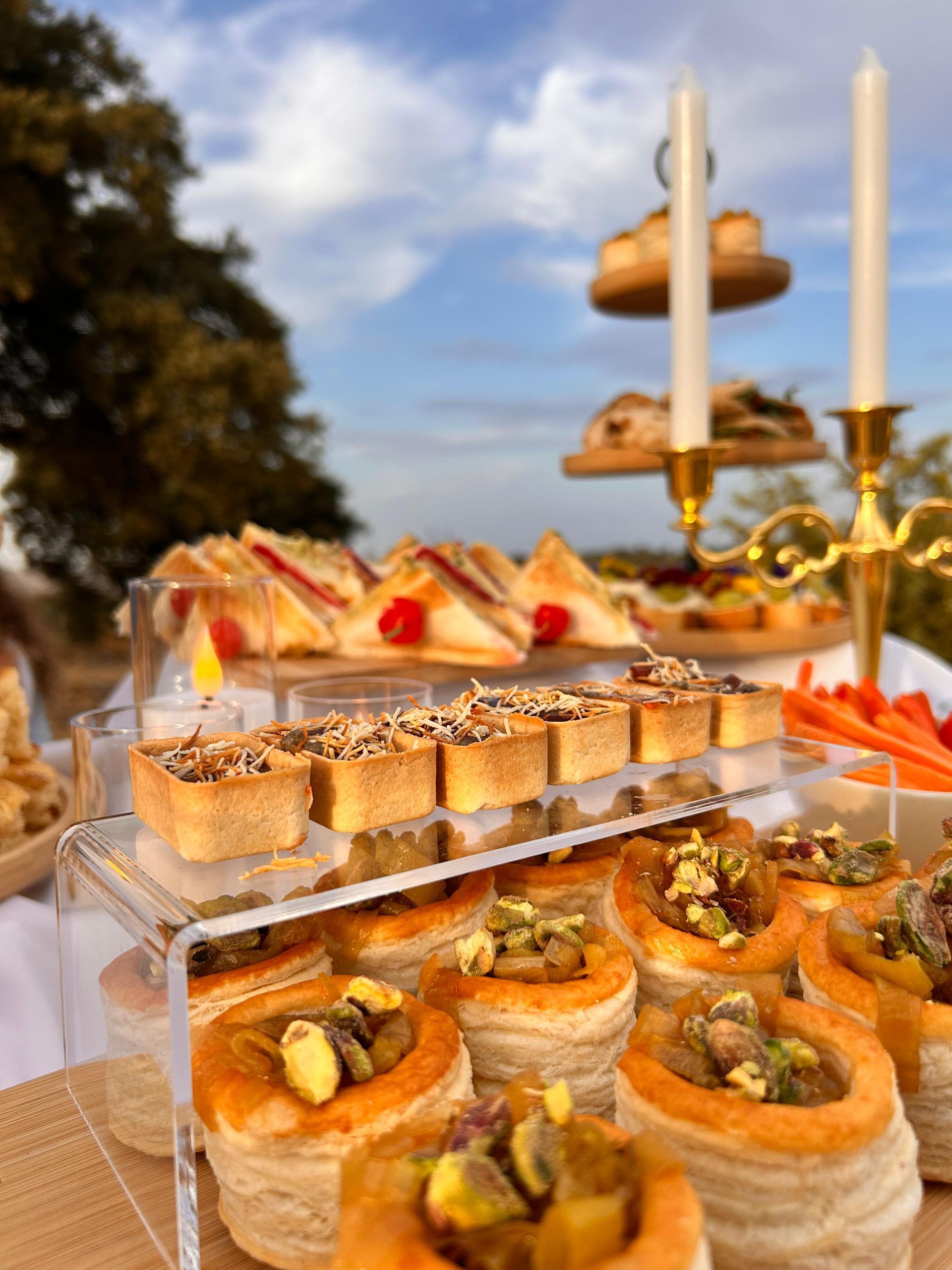 Bufé al aire libre: pasteles, sándwiches y aperitivos en una mesa blanca con un puesto de postres escalonado. Cielo azul.