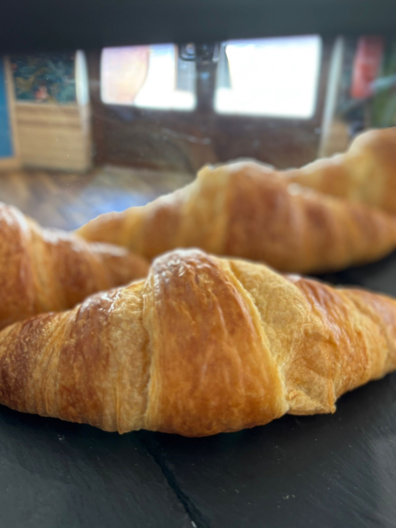 Croissants dorados sobre una superficie oscura, con un fondo borroso de una puerta y cajas.