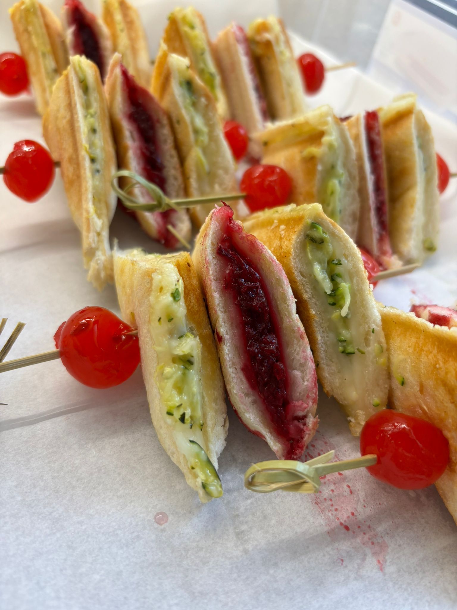 Sándwiches triangulares, ensartados con tomates cherry. Se ven varios rellenos: verde, rojo y blanco.