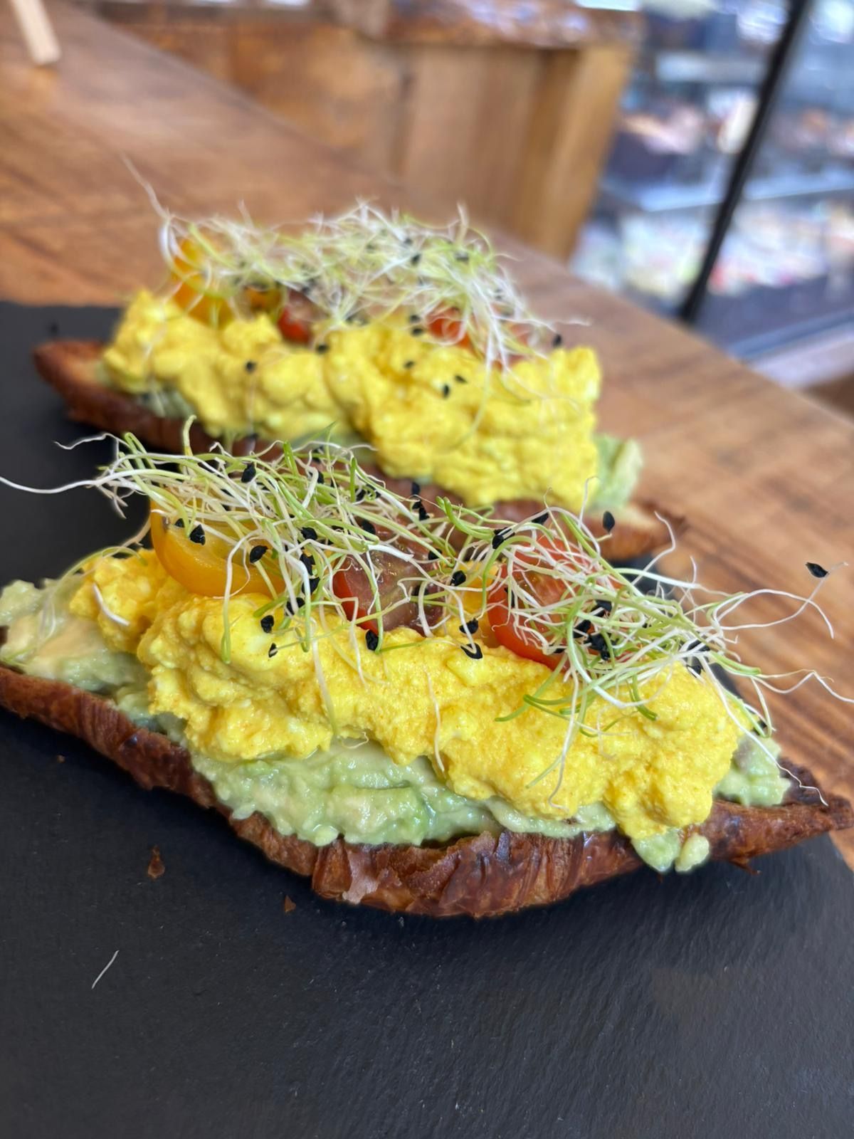Tostada de aguacate con tofu revuelto amarillo, brotes y semillas negras sobre una superficie oscura.