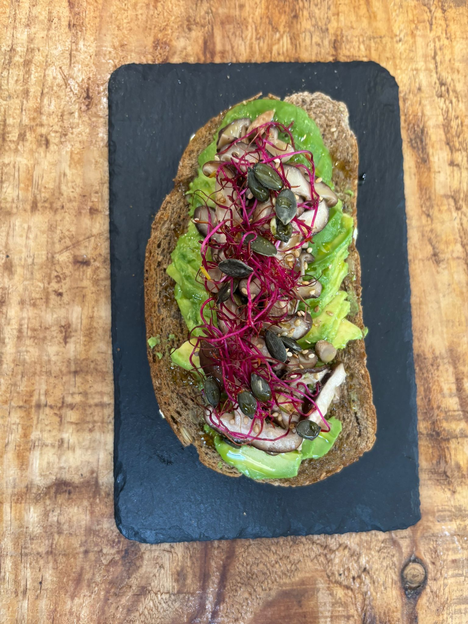 Tostada de aguacate sobre pan oscuro, cubierta con semillas, brotes y champiñones sobre una pizarra negra.