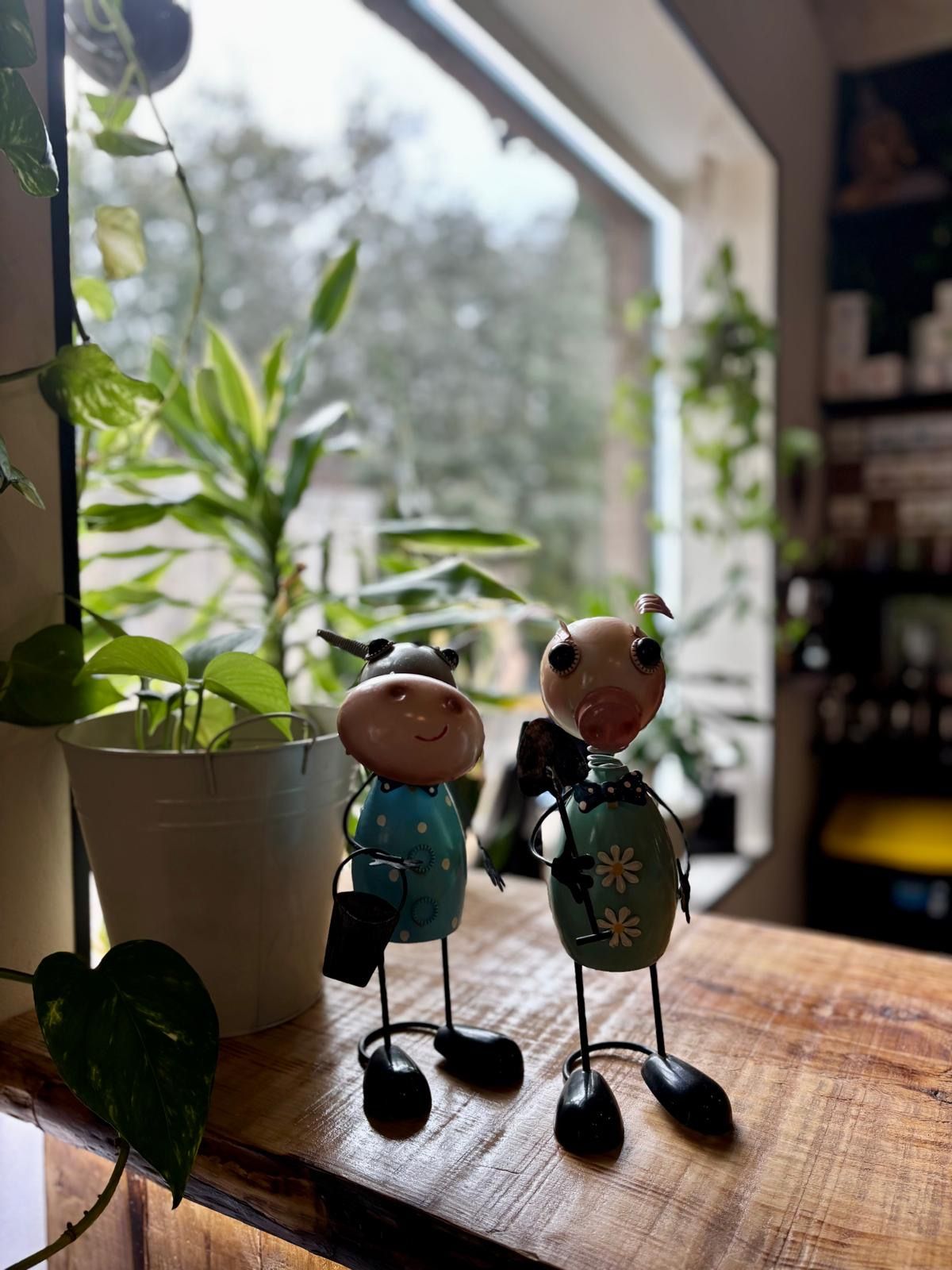 Dos figuras decorativas de metal se encuentran sobre una repisa de madera cerca de una ventana con plantas.