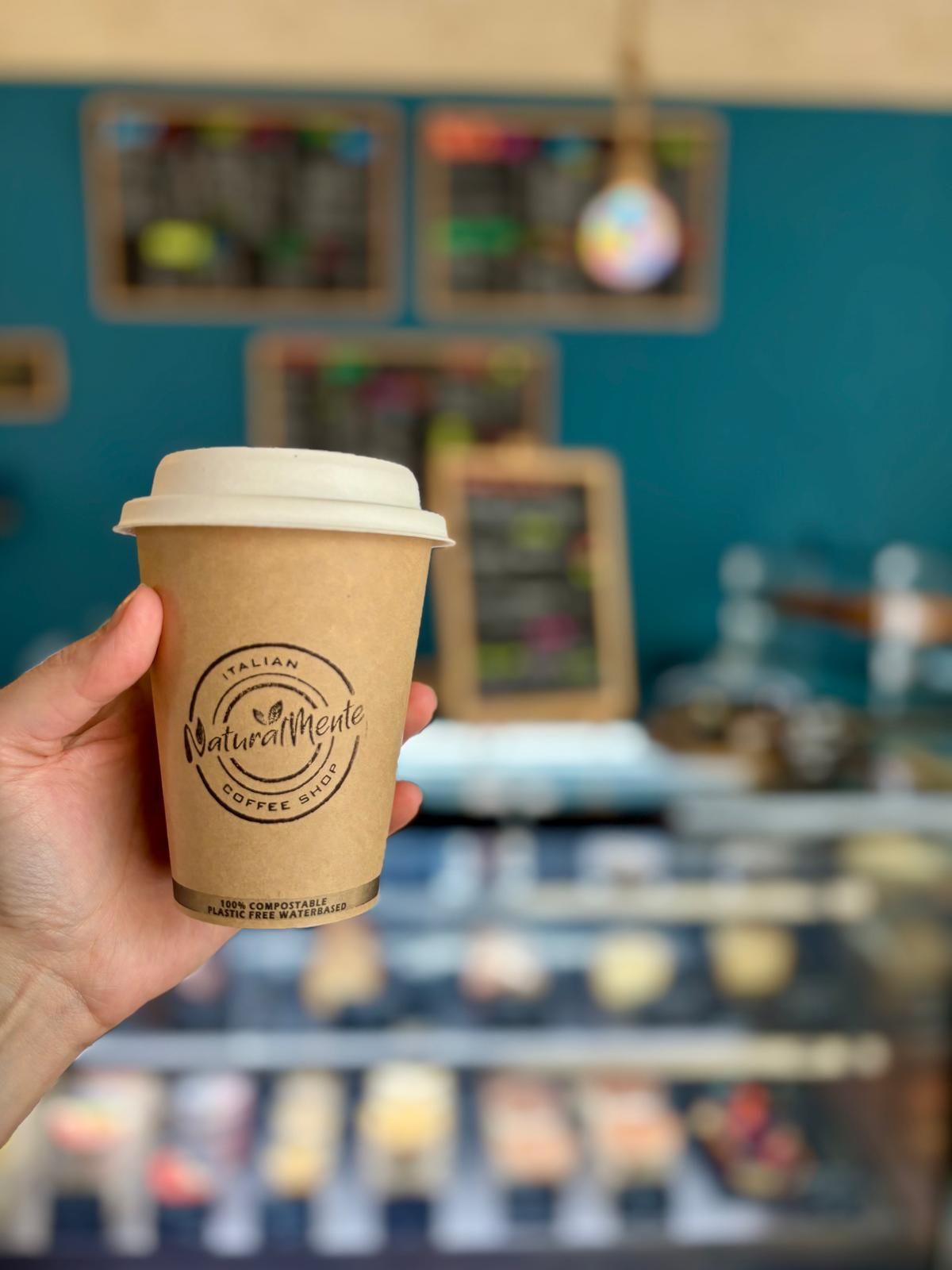 Mano sosteniendo una taza de café con el logo de una cafetería, frente a una pared turquesa y una vitrina de cafetería.