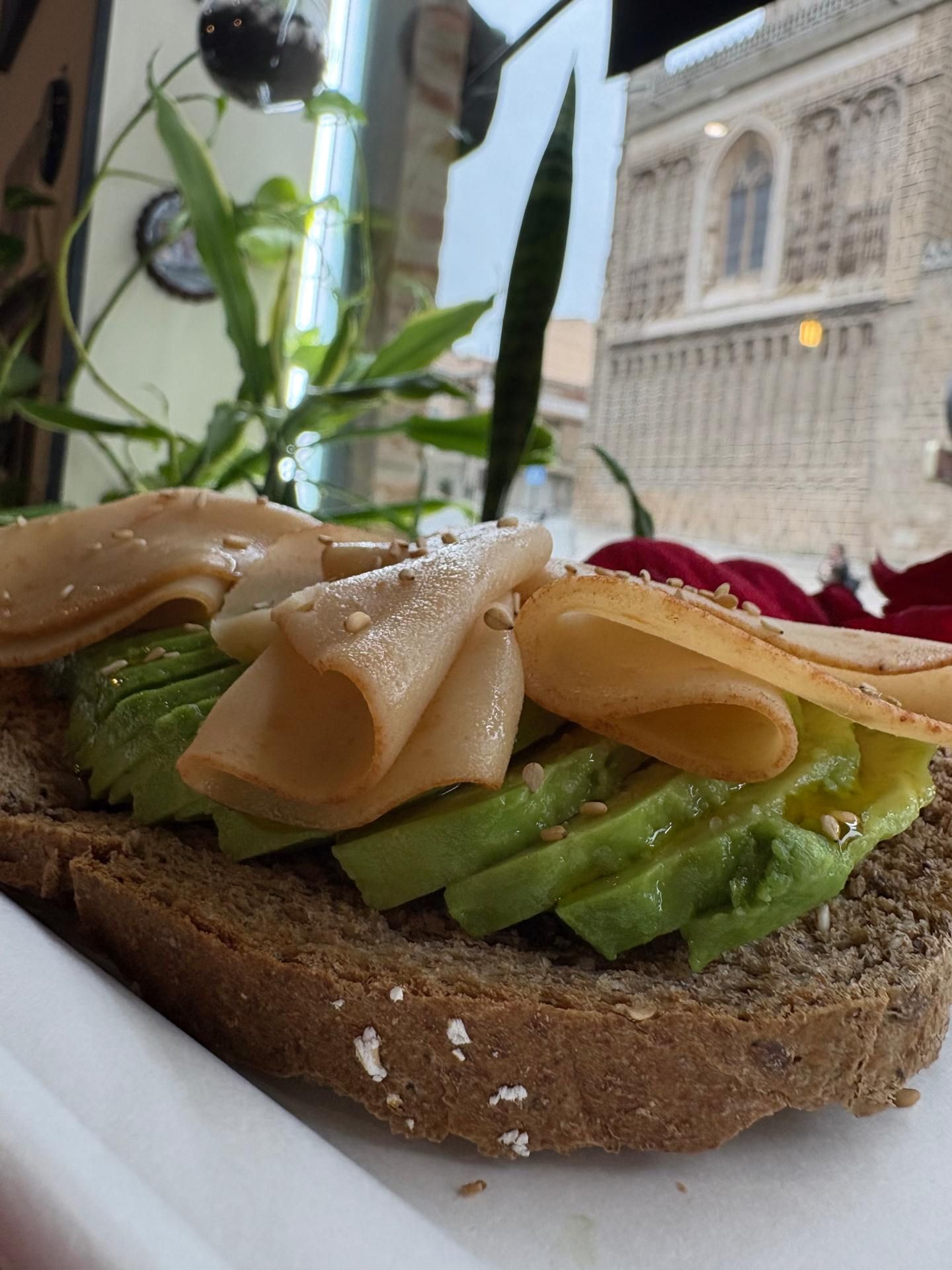 Tostada de aguacate con rodajas de pavo, semillas espolvoreadas y un edificio histórico de fondo.