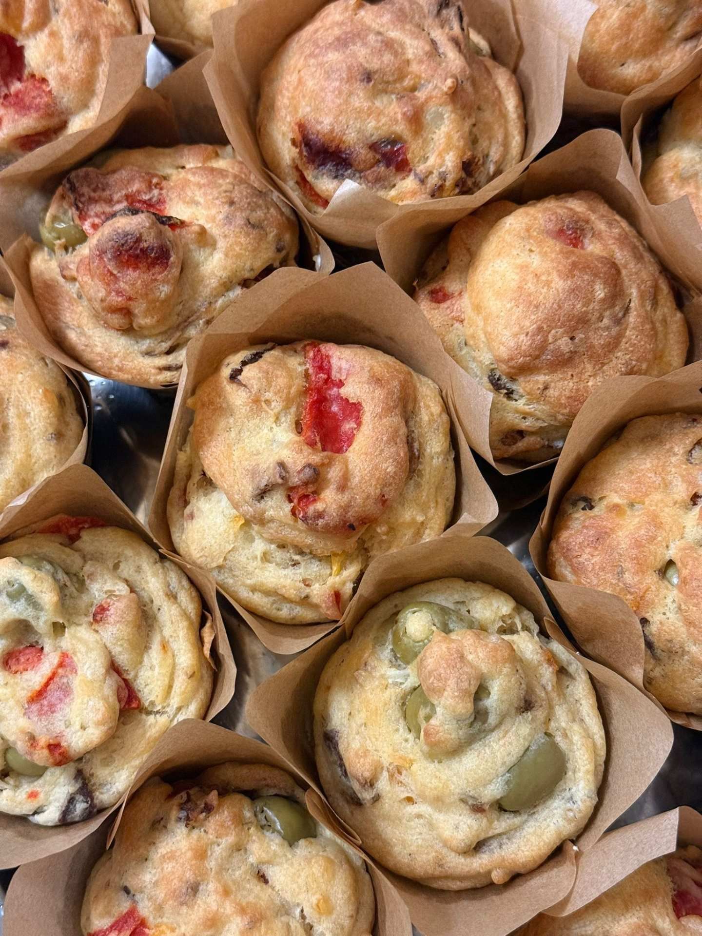 Muffins salados con aceitunas visibles y pimientos rojos envueltos en papel marrón.