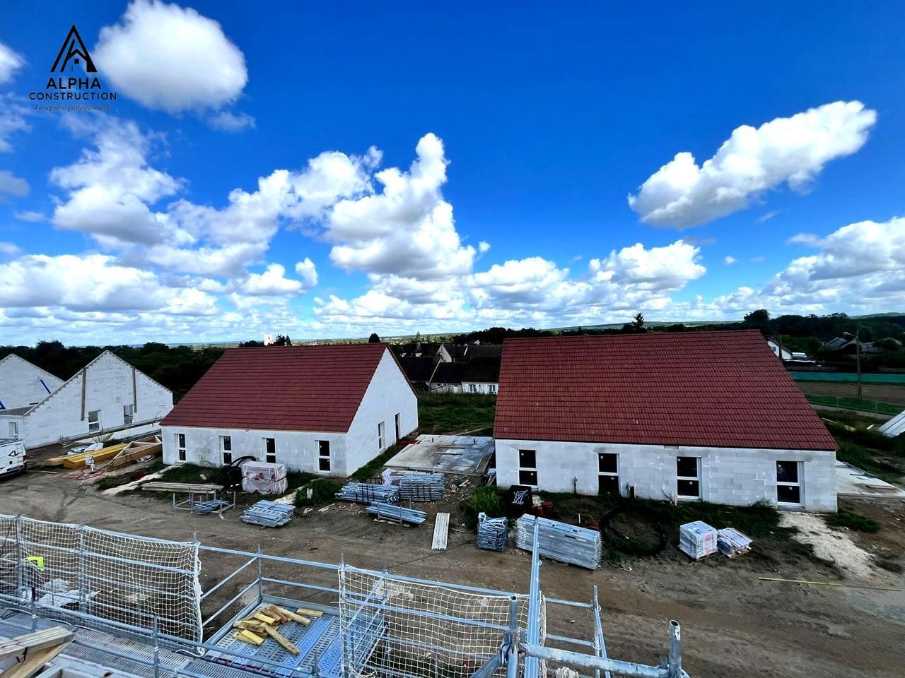 Deux maisons blanches aux toits rouges sous un ciel bleu avec des nuages ; chantier de construction.