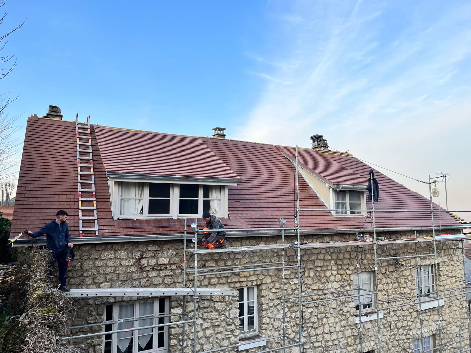 Des ouvriers sur un échafaudage réparent le toit d'une maison en pierre avec de nouvelles tuiles rouges sous un ciel bleu.