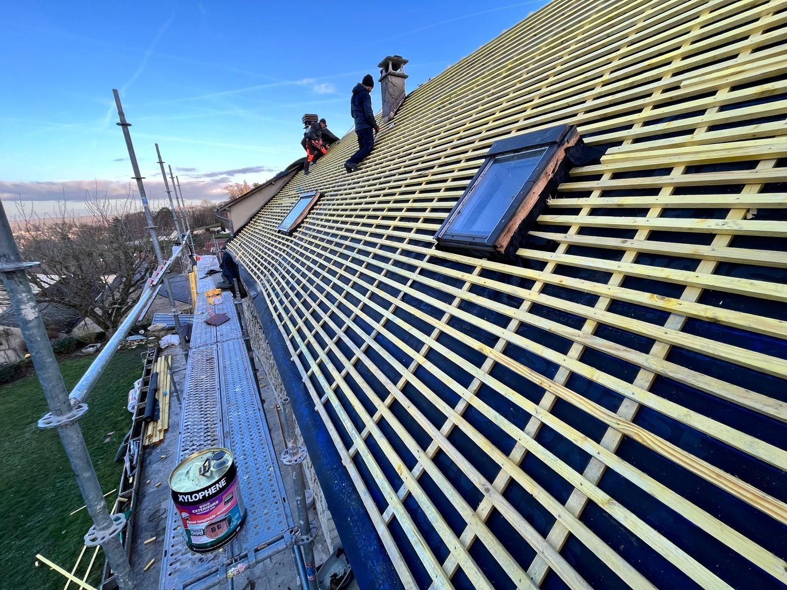 Des ouvriers installent des tasseaux de bois sur un toit. On aperçoit un échafaudage et une lucarne.