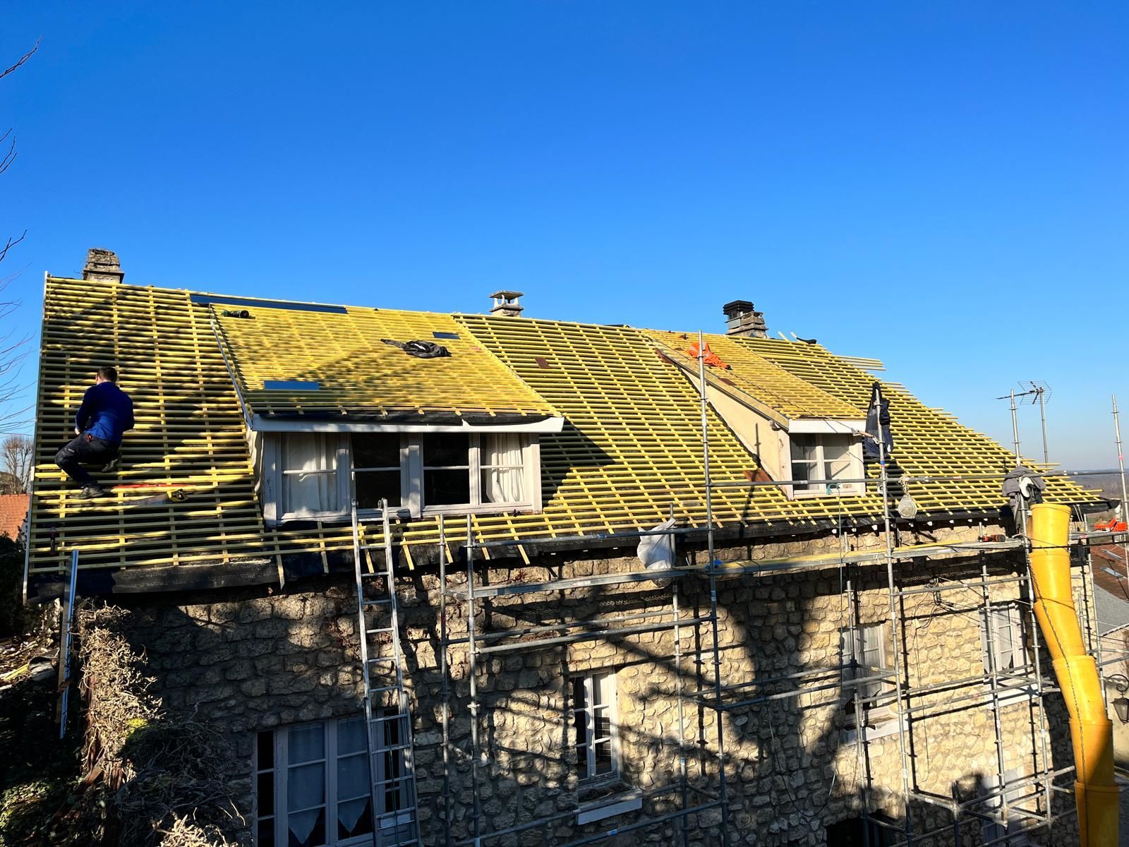 Toiture en rénovation, ouvrier sur un toit à ossature bois. Ciel bleu, bâtiment en pierre clair.