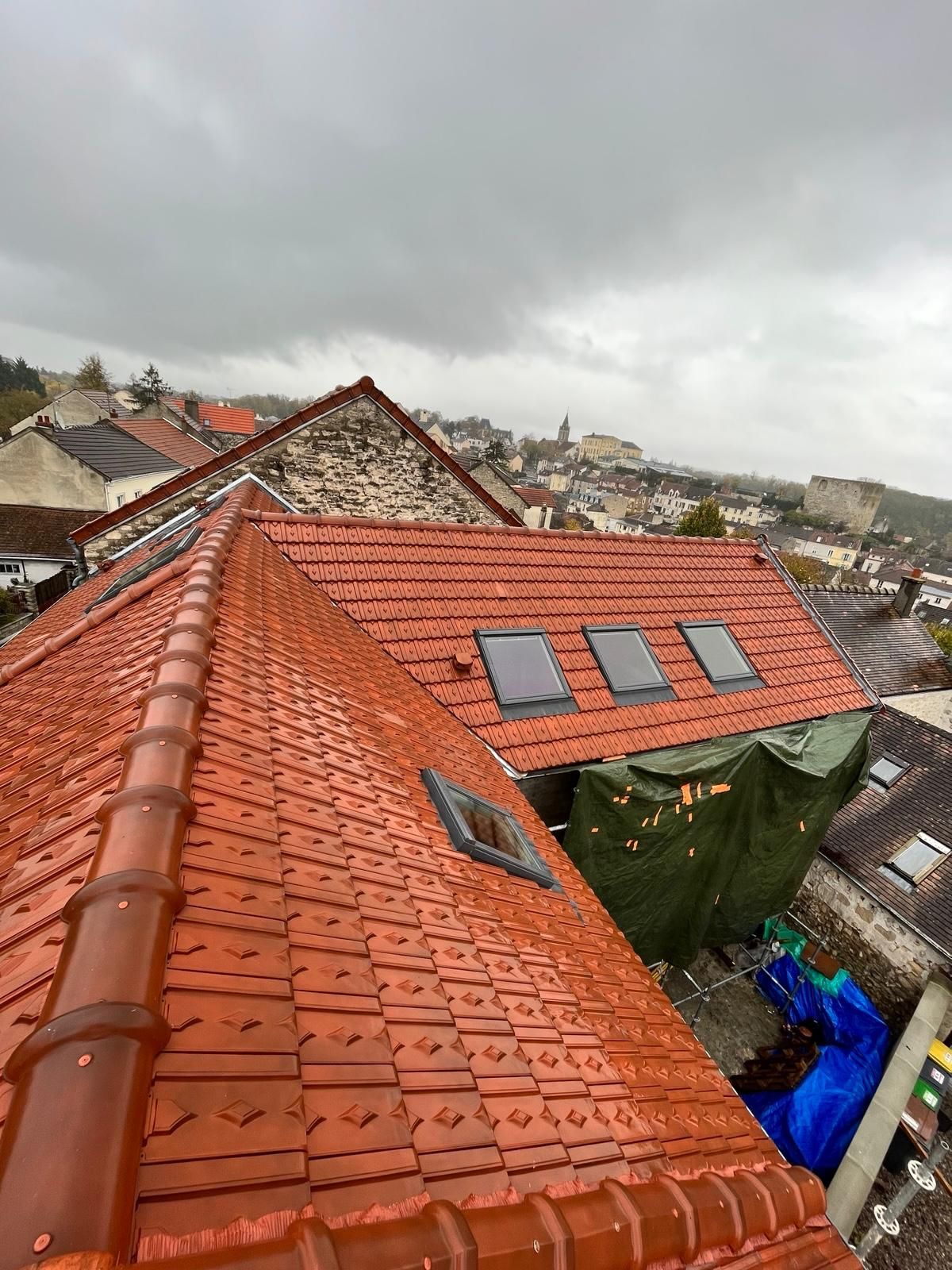 Toits de tuiles rouges, puits de lumière et paysage urbain environnant sous un ciel couvert.