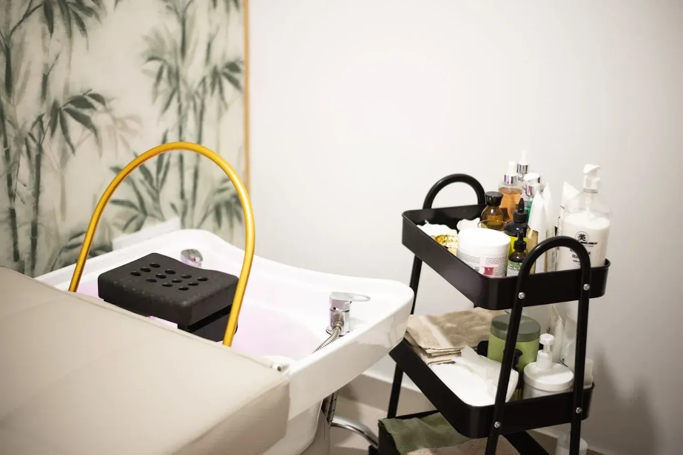 Un lavacabezas de salón con lavabo blanco, soporte curvo dorado y un carrito negro de varios niveles con productos de belleza.