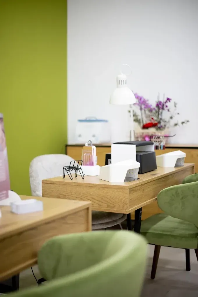 Un puesto de manicura en un salón de belleza con escritorios de madera, sillas de terciopelo verde y una lámpara blanca sobre una pared verde.